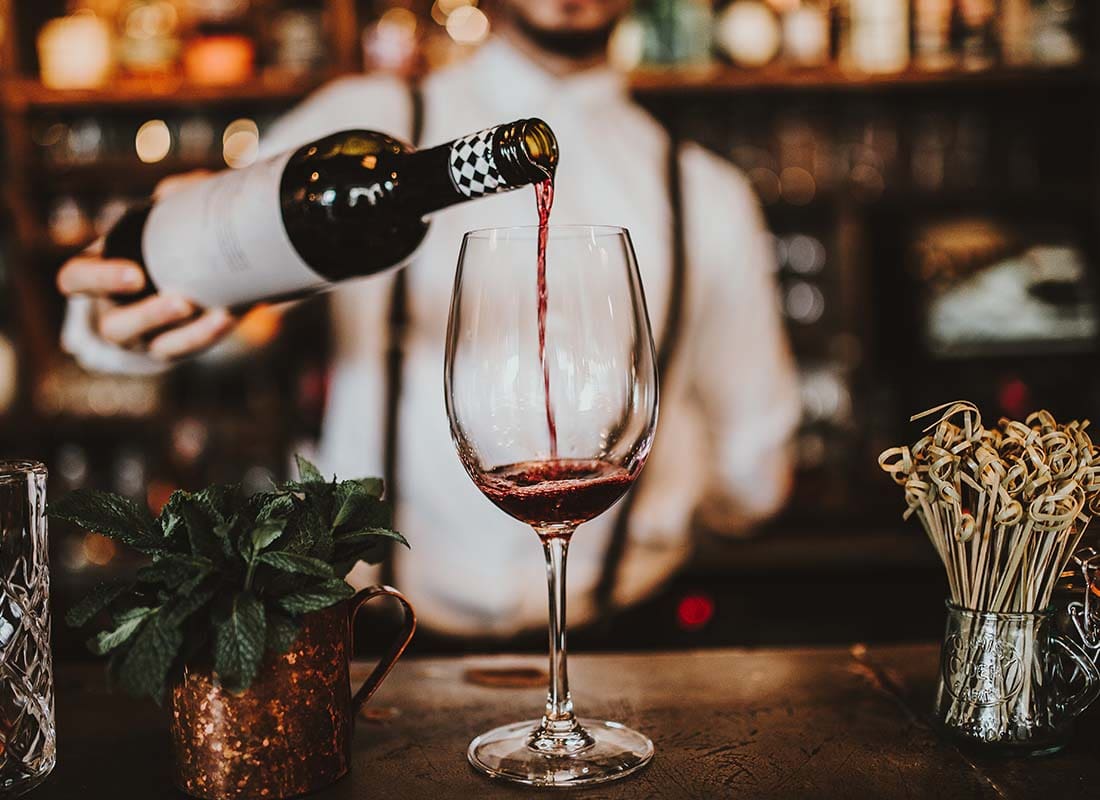 Liquor Liability Insurance - Close-up View of a Bartender Pouring Red Wine into a Glass over an Antique Bar with Bottles Blurred in the Distance